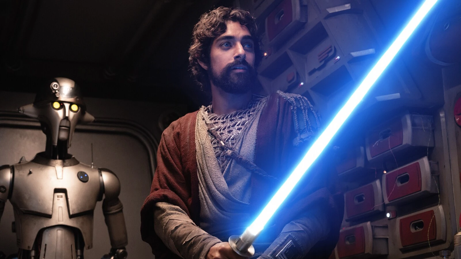 Ezra Bridger holds a blue lightsaber beside the droid Huyang in Star Wars: Ahsoka.