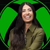Xbox CEO Asha Sharma smiles in front of a large green Xbox sphere logo.