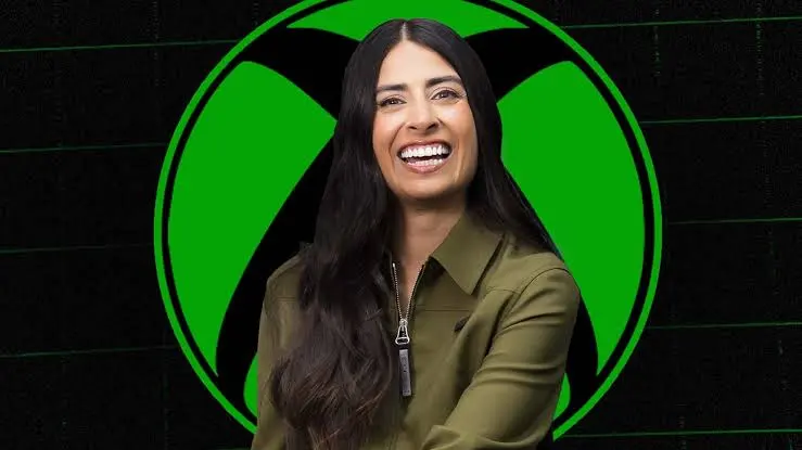 Xbox CEO Asha Sharma smiles in front of a large green Xbox sphere logo.