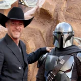 Brendan Wayne, in a black cowboy hat and suit, stands beside a costumed Din Djarin and Grogu at an event.