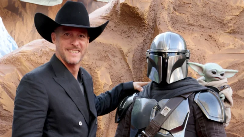 Brendan Wayne, in a black cowboy hat and suit, stands beside a costumed Din Djarin and Grogu at an event.