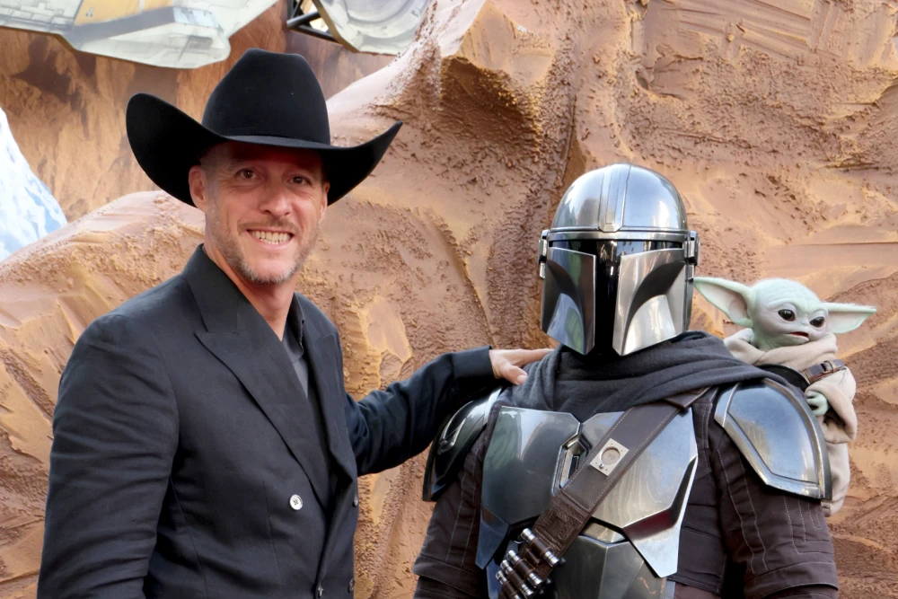 Brendan Wayne, in a black cowboy hat and suit, stands beside a costumed Din Djarin and Grogu at an event.