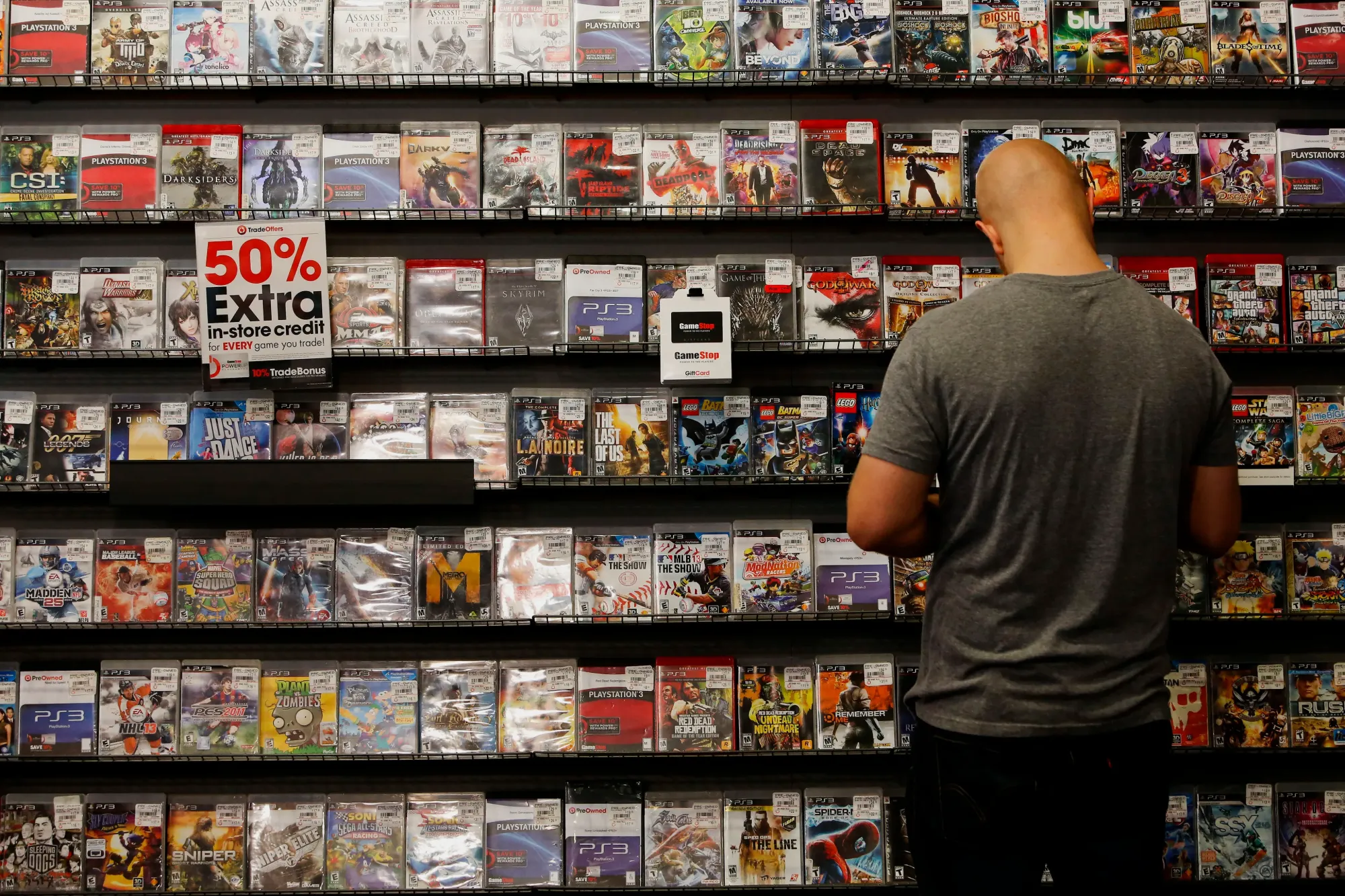 A customer browses a wall of pre-owned PlayStation 3 games at a GameStop beside a 50 percent extra trade-in credit sign.