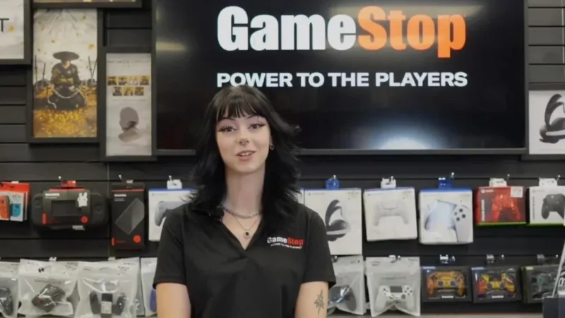 A GameStop store associate in a black polo stands in front of a Power to the Players wall of controllers.