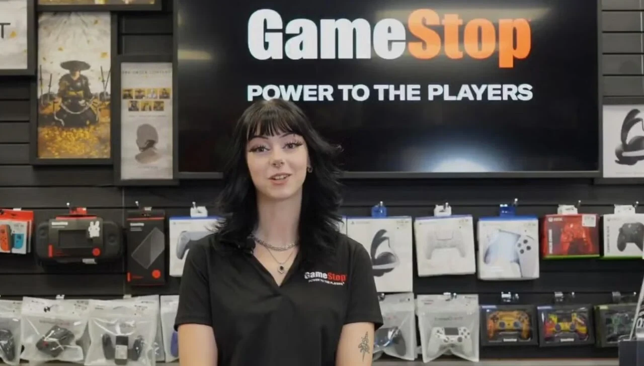 A GameStop store associate in a black polo stands in front of a Power to the Players wall of controllers.