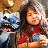 Stitch grins beside Lilo, played by Maia Kealoha, in a sunlit scene from Disney’s live-action Lilo & Stitch.