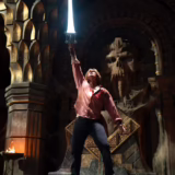 Prince Adam raises the glowing Power Sword in a torchlit throne room beneath a giant carved skull in Masters of the Universe.
