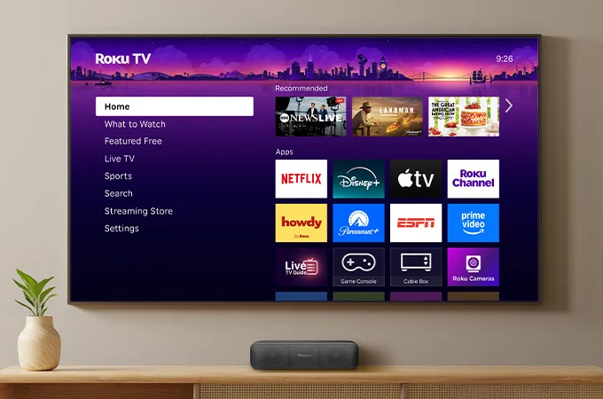 A Roku TV home screen on a wall-mounted television showing streaming apps including Netflix, Disney+, and Tubi.
