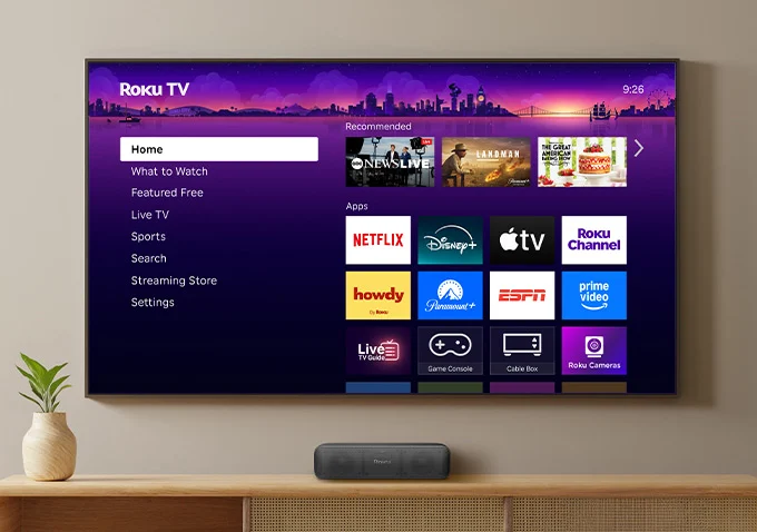 A Roku TV home screen on a wall-mounted television showing streaming apps including Netflix, Disney+, and Tubi.